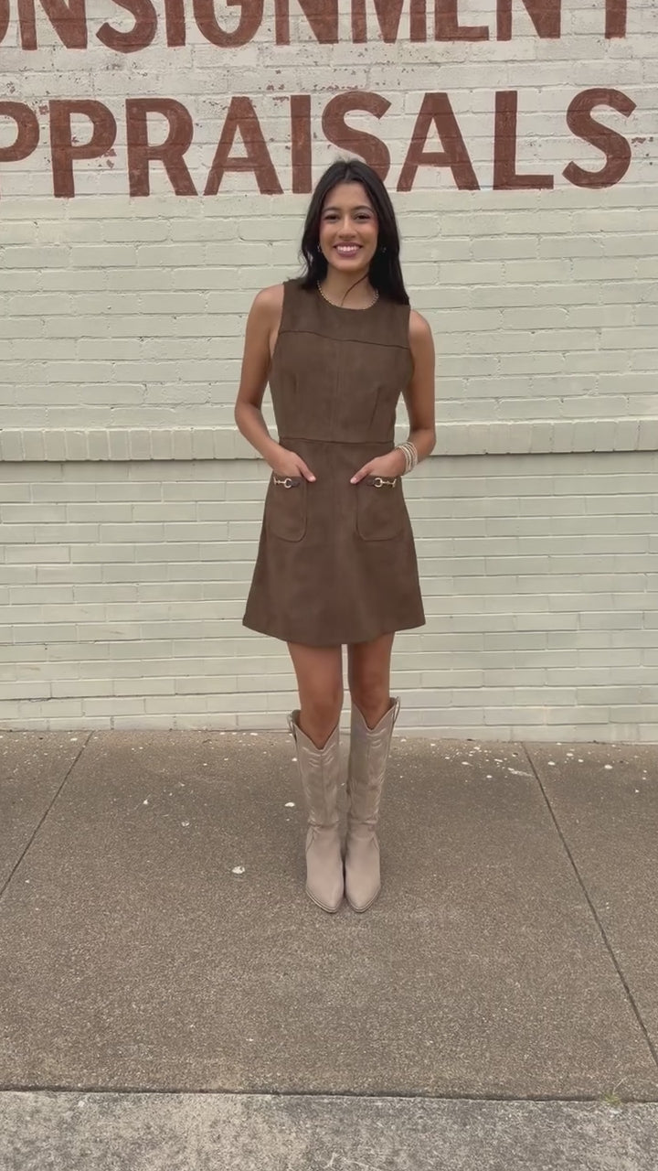 Model wearing the Sorrel Mini Dress in mocha with a suede-like finish, sleeveless design, gold horsebit pocket accents, and cream western boots. Southern House Clothing