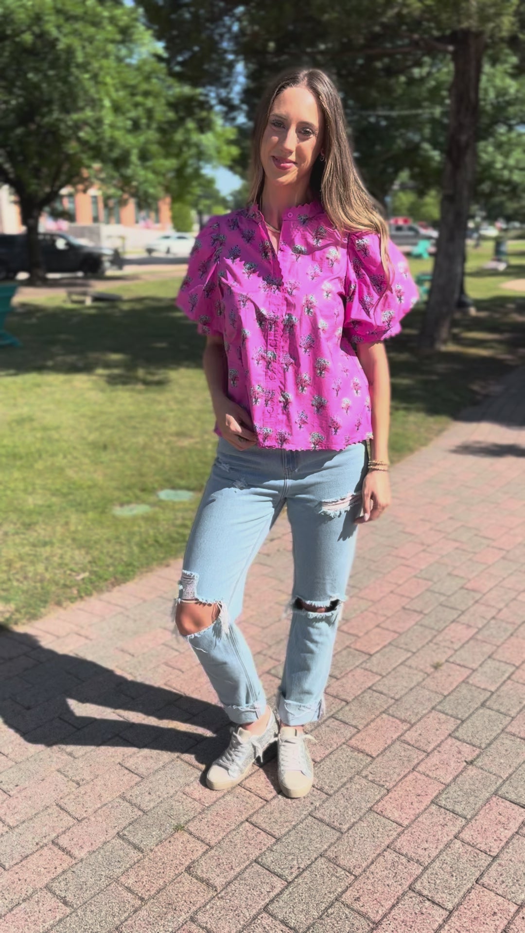A woman wearing a pink short-sleeve button-up shirt with a black palm tree print, light-wash distressed jeans, and white sneakers, walking outdoors.