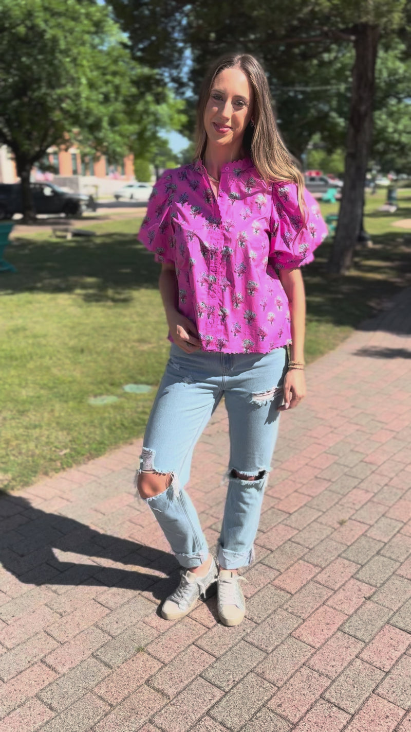 A woman wearing a pink short-sleeve button-up shirt with a black palm tree print, light-wash distressed jeans, and white sneakers, walking outdoors.