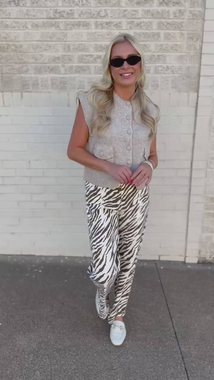A woman modeling a light gray sleeveless button-front vest and wide-leg zebra-print pants, standing outdoors against a white brick wall.