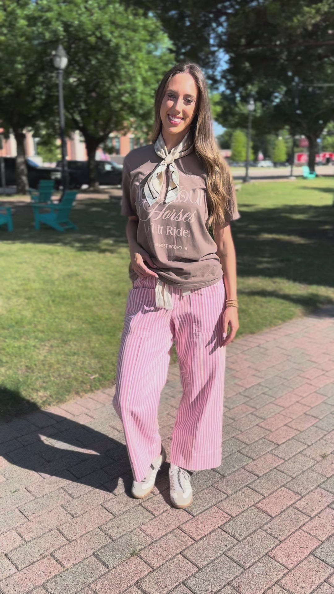 A woman stands outdoors, wearing a brown graphic T-shirt, pink and white vertically striped wide-leg pants, a cream neck scarf, and white sneakers.