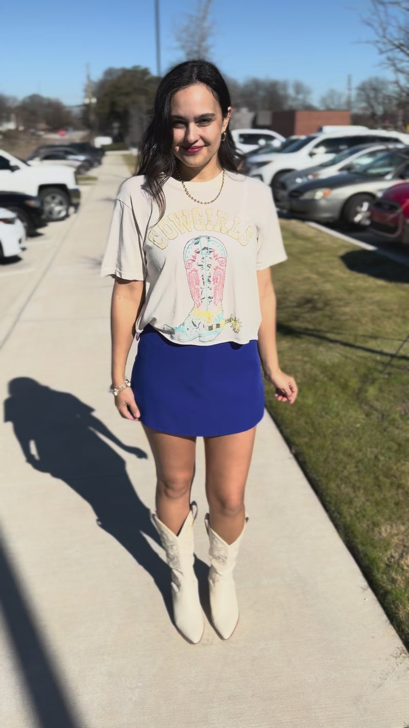 Woman wearing a graphic t-shirt and blue skirt against outside.