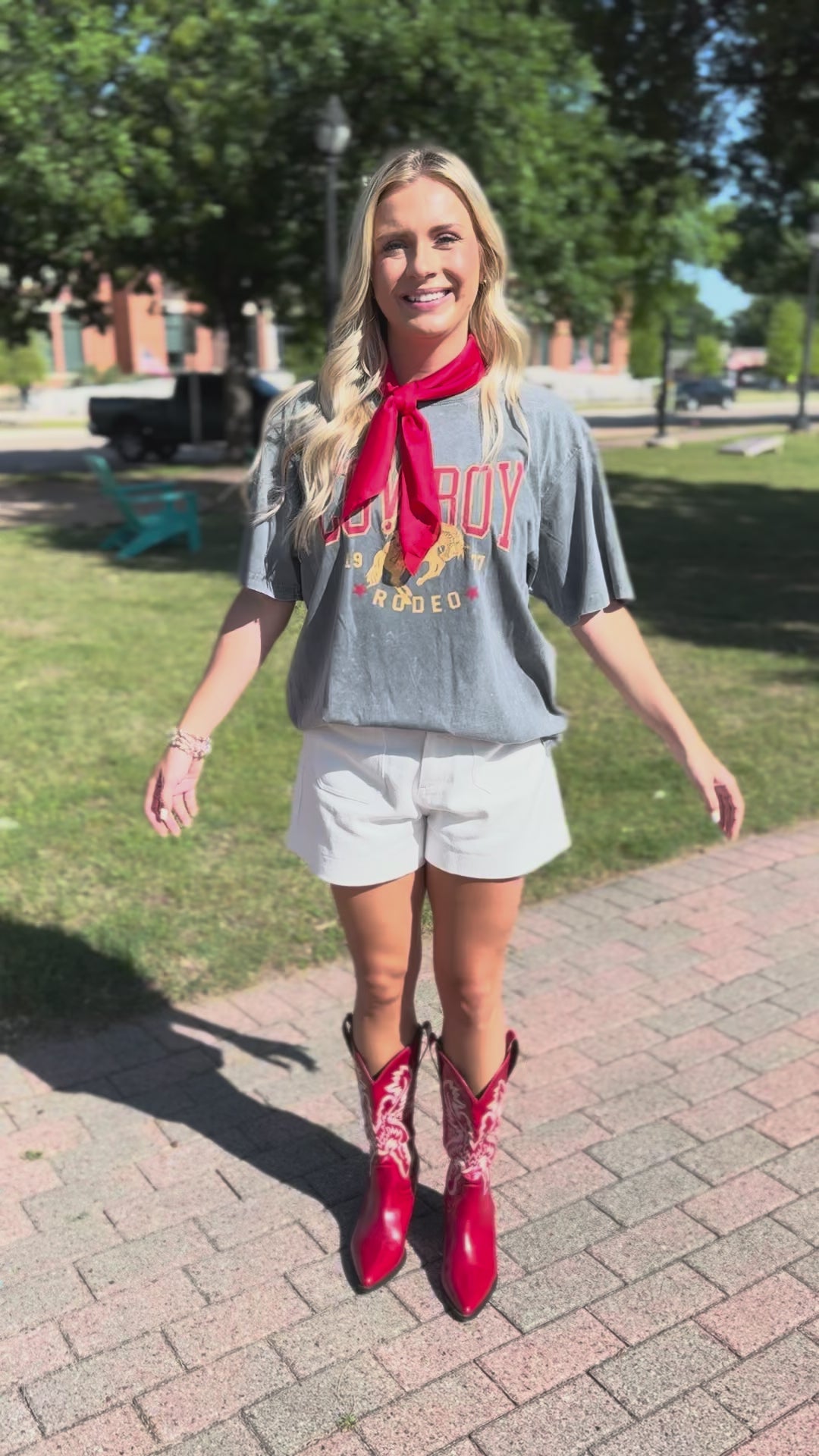 A woman posing on a city sidewalk wearing a gray graphic t-shirt with a red neck scarf, white shorts, and shiny red cowboy boots.