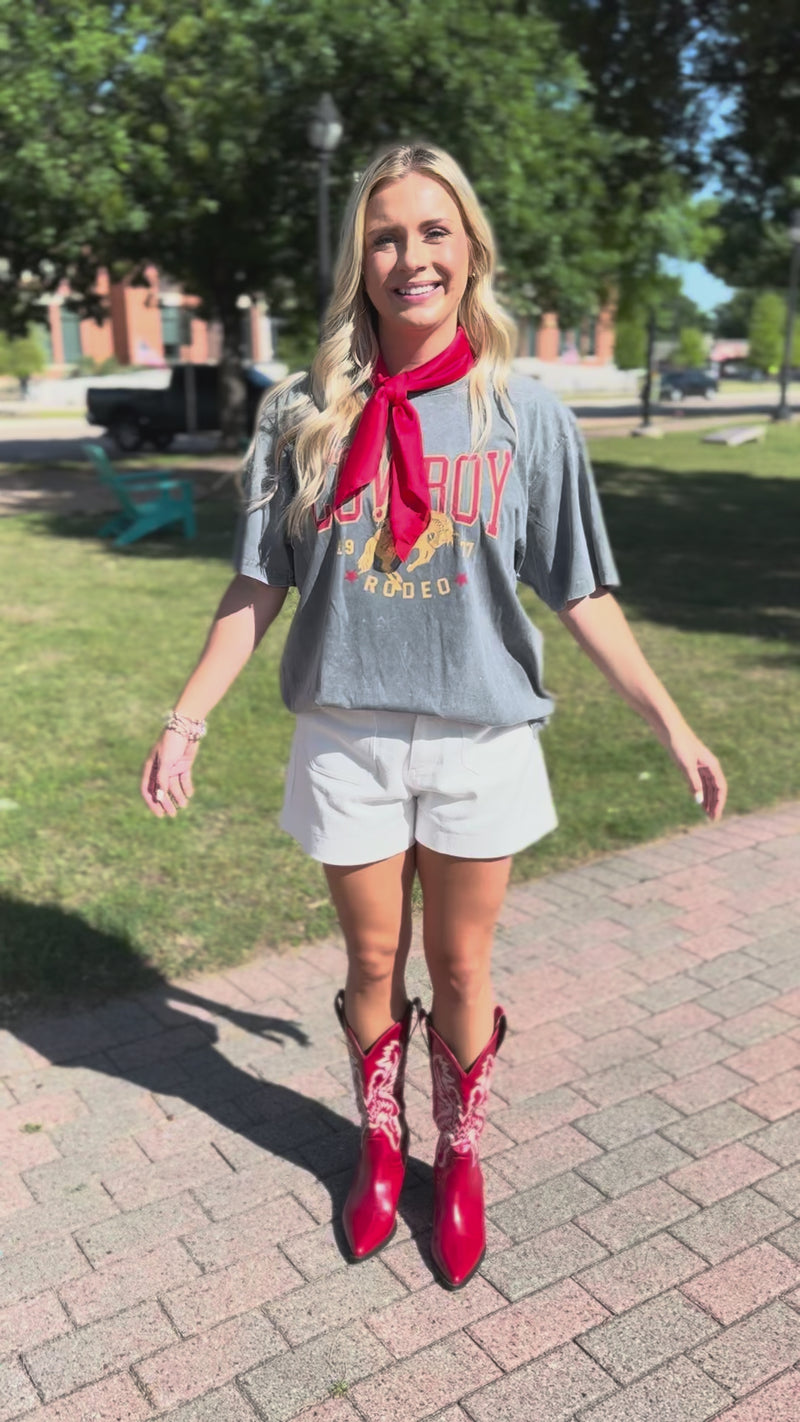 A woman posing on a city sidewalk wearing a gray graphic t-shirt with a red neck scarf, white shorts, and shiny red cowboy boots.