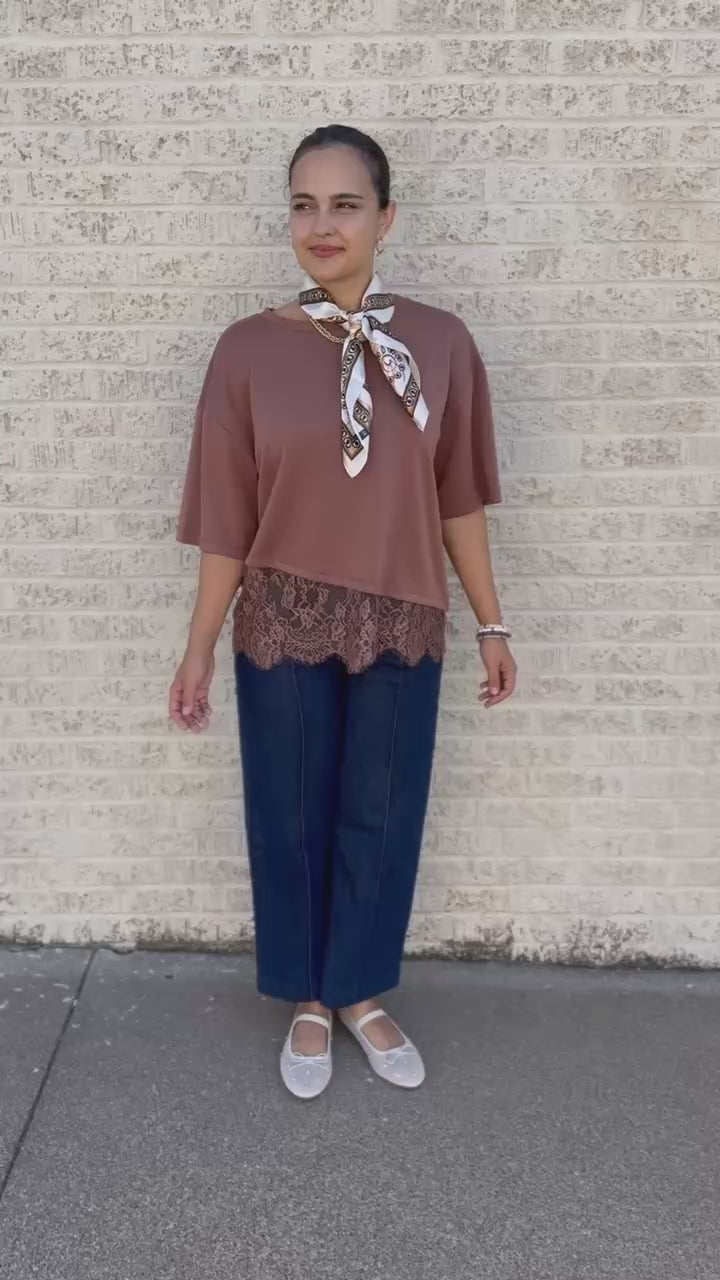 A woman stands on a sidewalk wearing a dusty rose short-sleeve top with a lace hem, a patterned neck scarf, dark blue wide-leg pants, and white flats, holding a small sage green handbag.