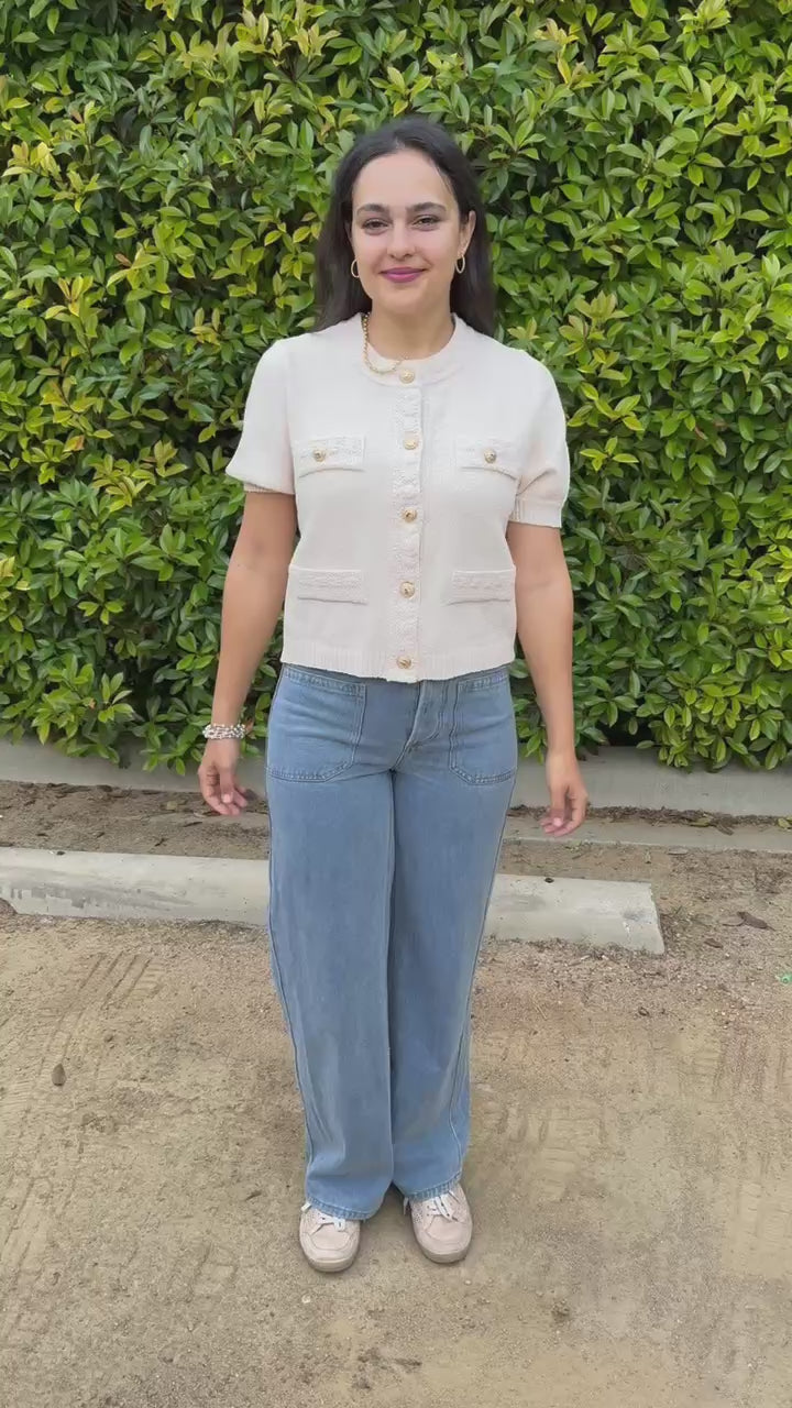 Woman in a beige cardigan and blue jeans standing outside.