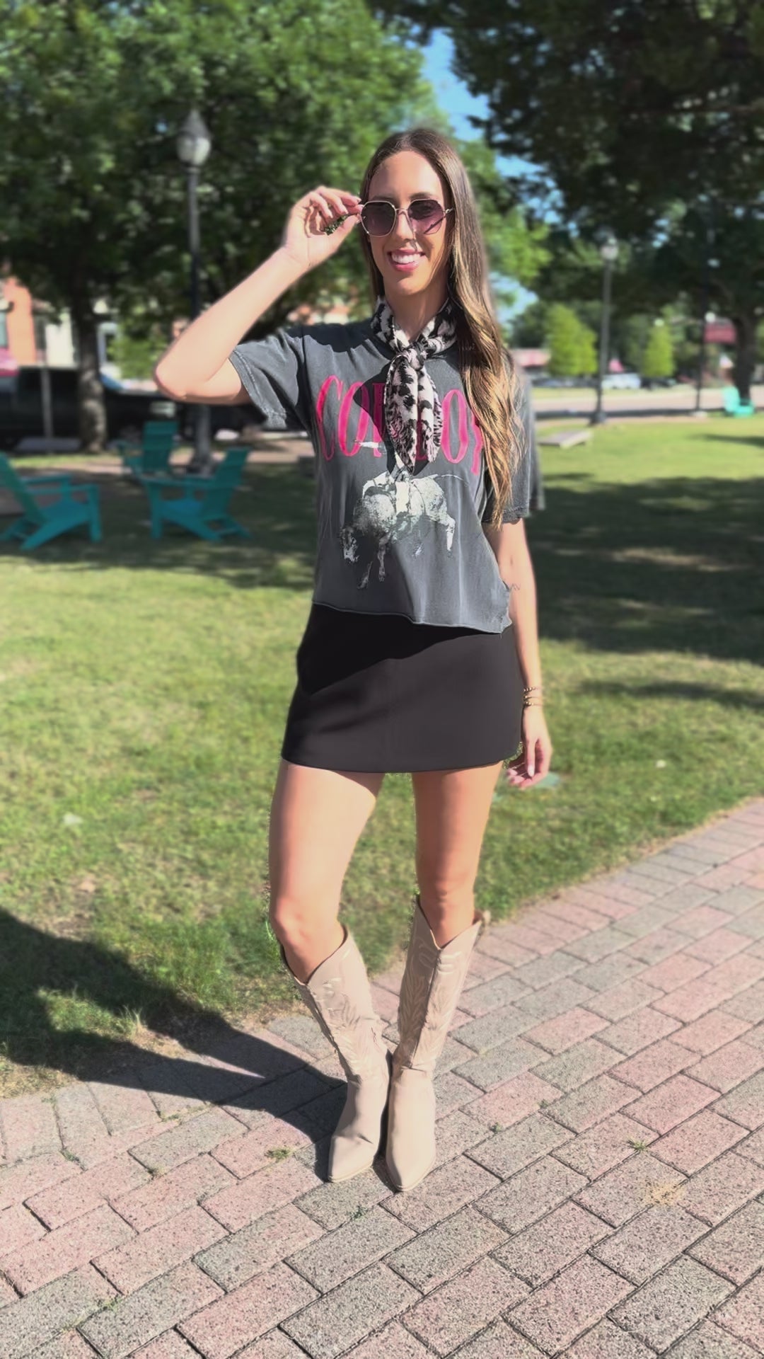 Woman standing outside wearing a black graphic tee, scarf on her neck, black skirt, and tan boots.