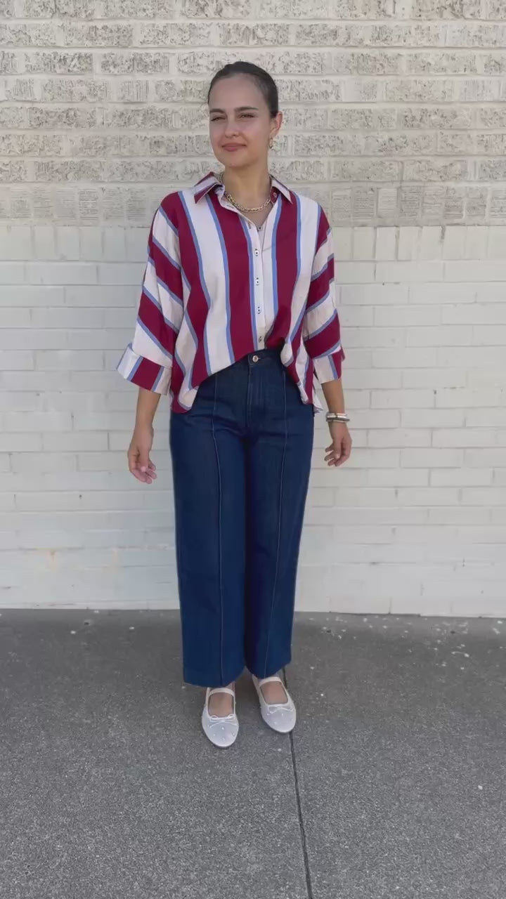 A woman stands outdoors against a light brick wall wearing a red, white, and blue vertically striped button-up shirt, dark blue wide-leg jeans, and white flats.