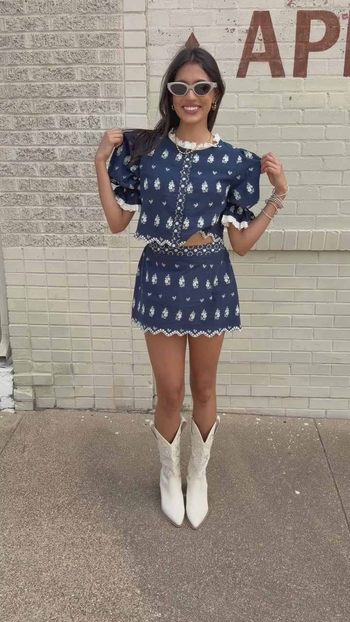 Brunette in a blue set standing outside wearing white cowboy boots and sunglasses. Southern House Clothing