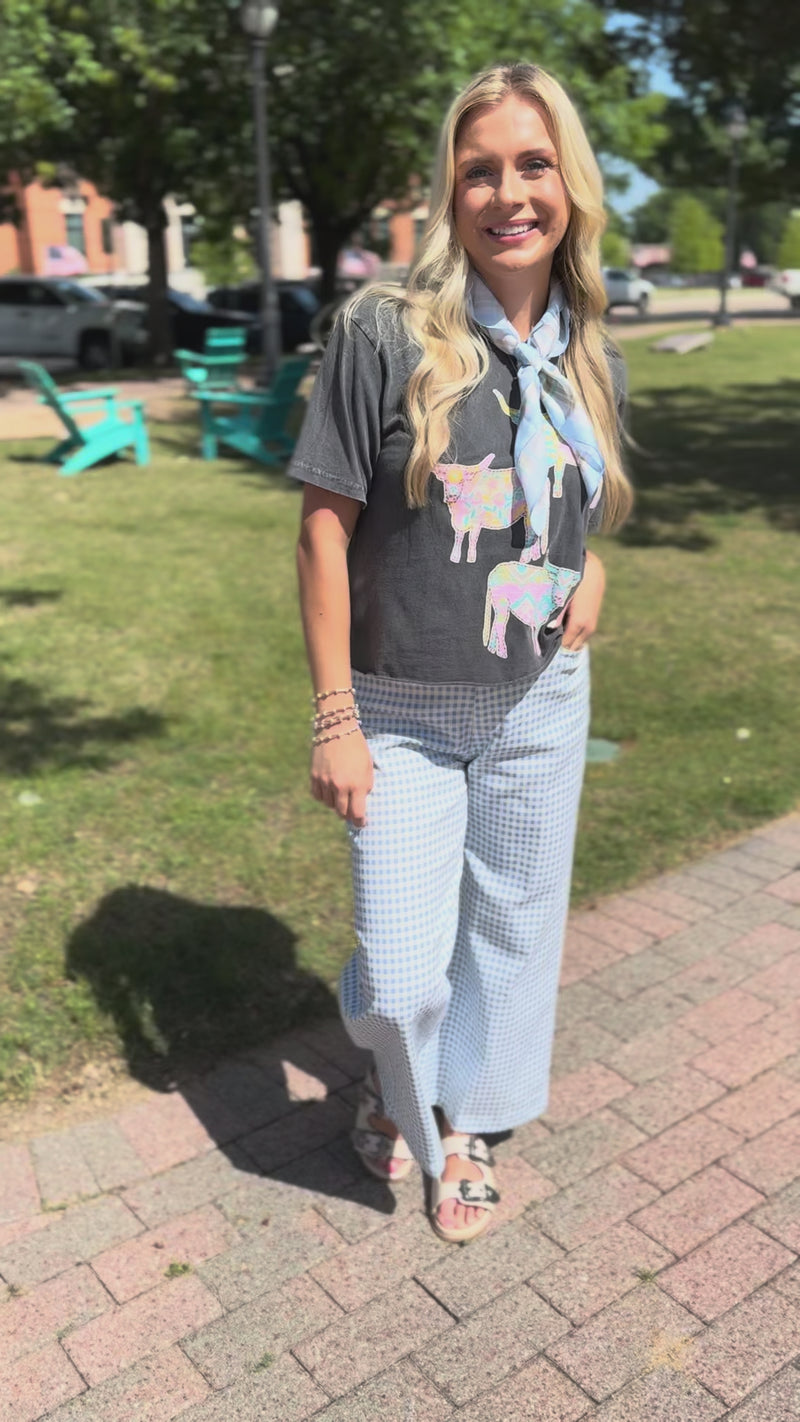 Blonde model wearing a black graphic tee, a blue scarf, and blue gingham pants, standing outside.