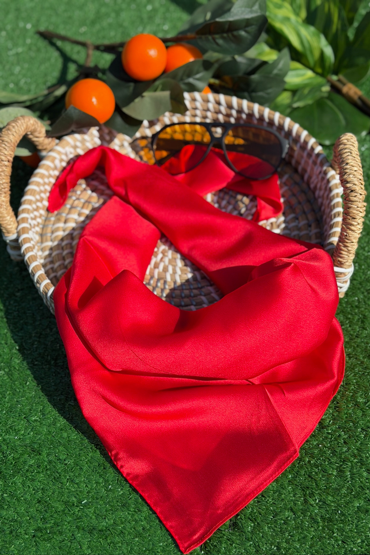 Woman's red scarf laying outside on a tan wicker basket with sunglasses and an orange floral arrangement. Margo scarf- red