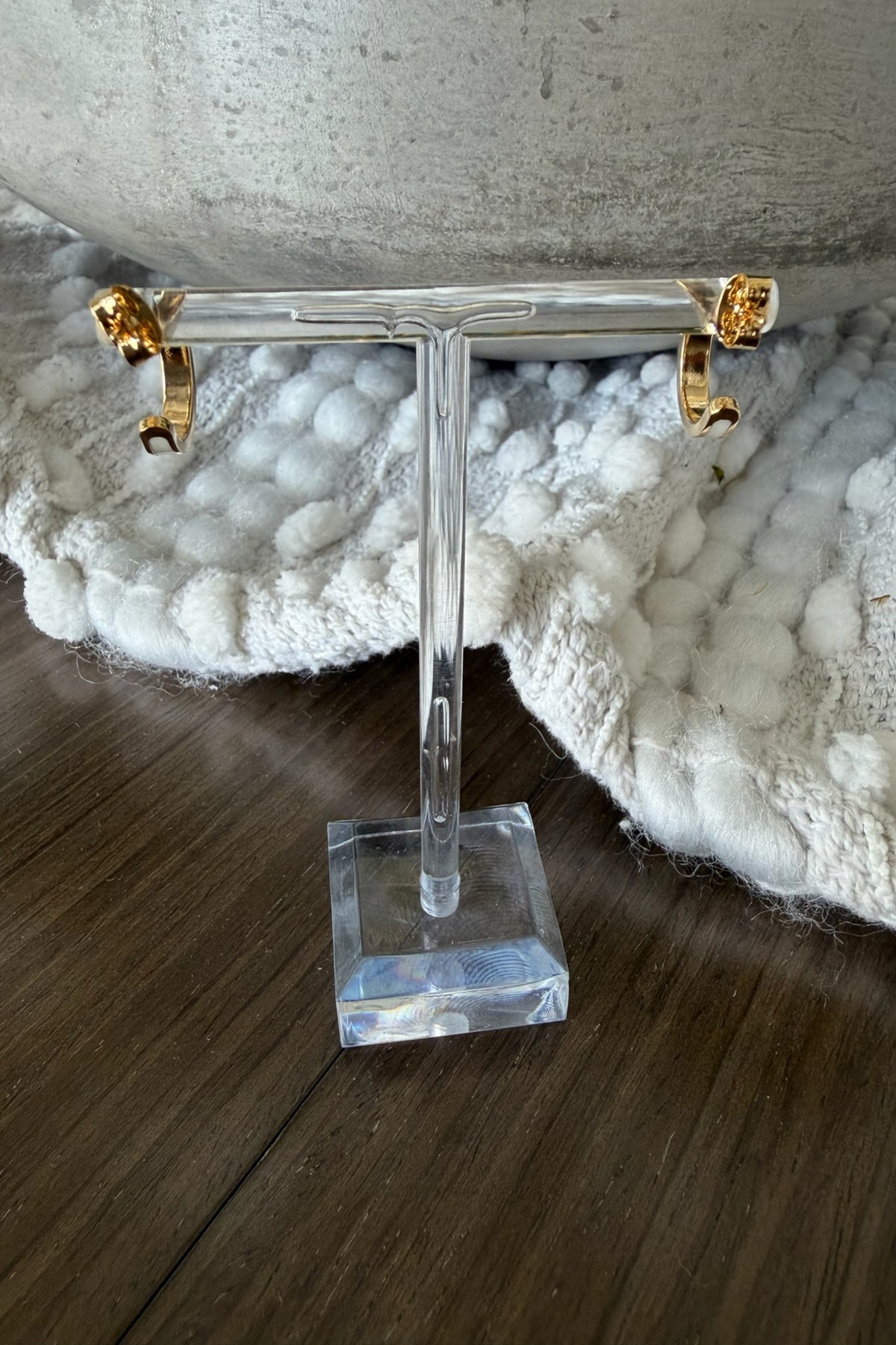 Gold hoop earrings on a clear T-shaped stand with a textured white surface and wooden floor in the background.