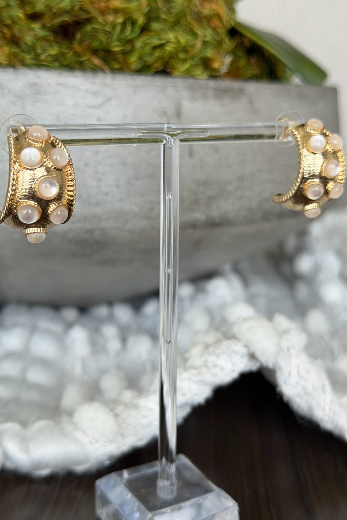 Gold earrings with pearl-like stones on a clear stand against a blurred natural background